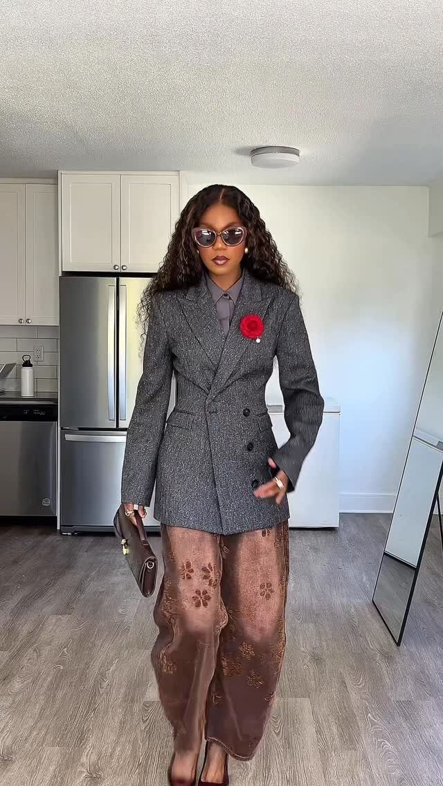 Woman in a grey blazer and patterned trousers wearing dark sunglasses and holding a handbag.