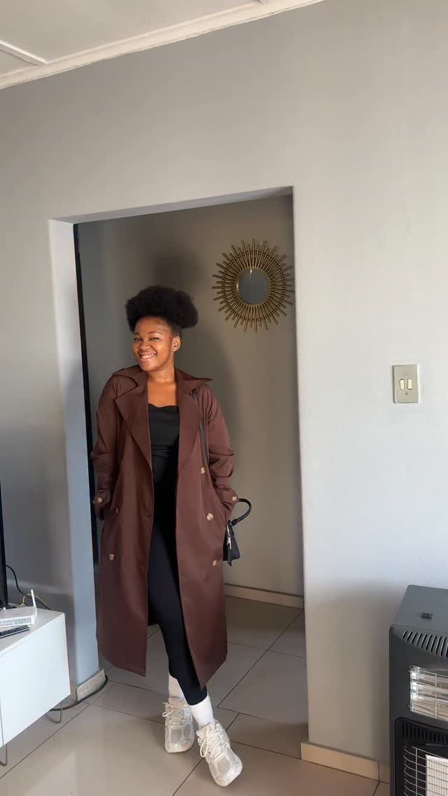15 Trench Coat Outfit Ideas for Dressing With Intention 10 A woman smiling in a brown trench coat, black top, and black leggings with white sneakers.