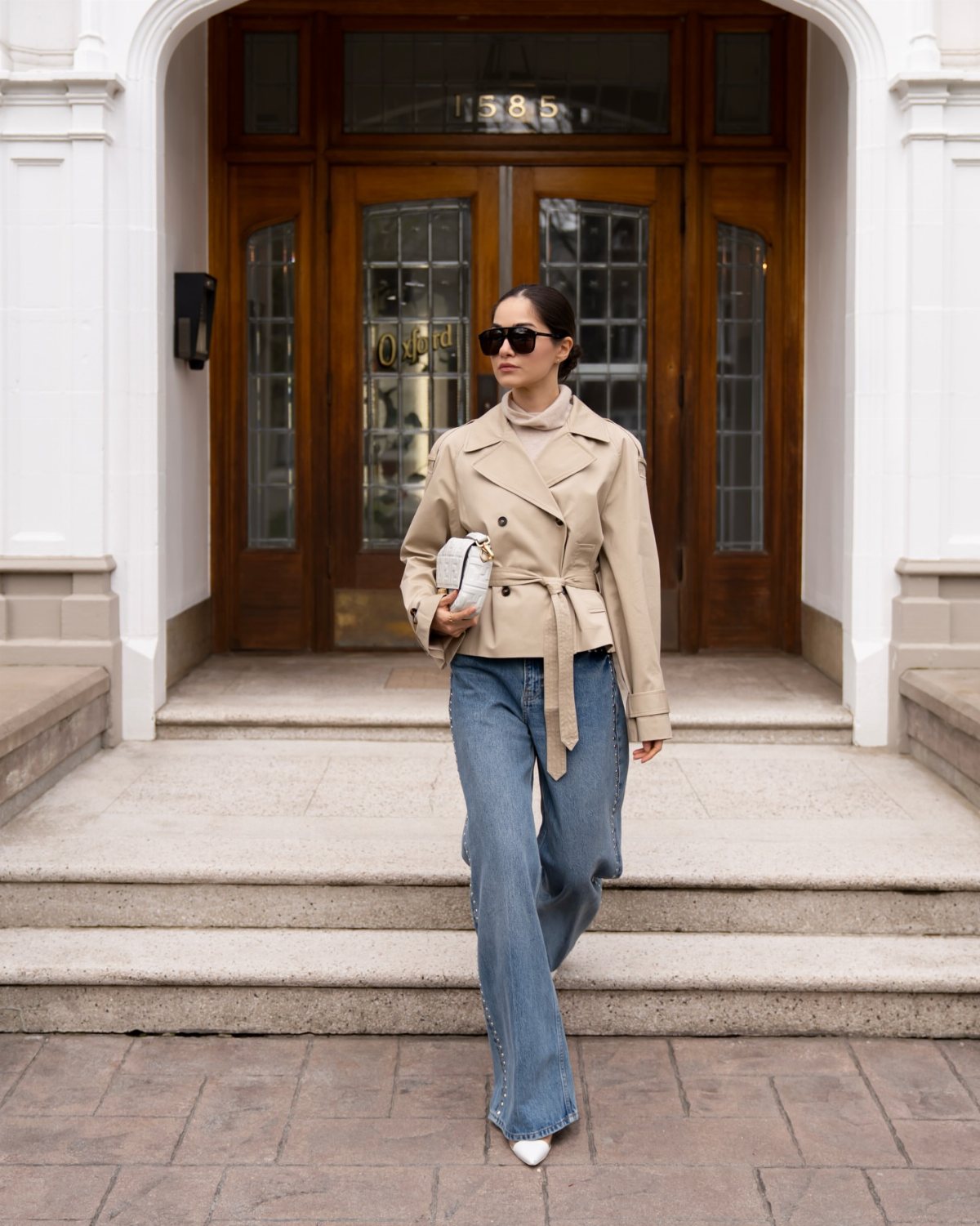 Woman in a cropped beige trench coat and blue wide-leg jeans standing on stone steps.