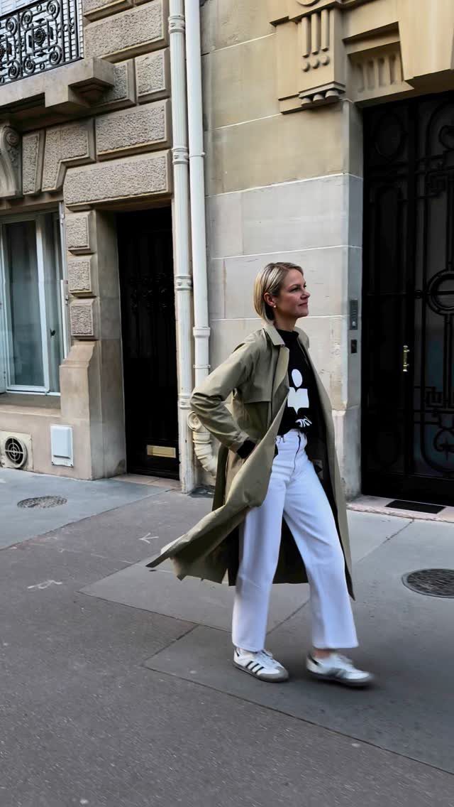 15 Trench Coat Outfit Ideas for Dressing With Intention 5 Woman in an olive trench coat, black graphic tee, white trousers, and white sneakers walking outdoors.