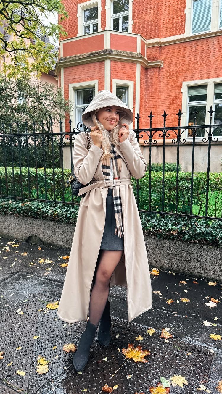 A woman in a long beige trench coat and bucket hat standing on a wet sidewalk.