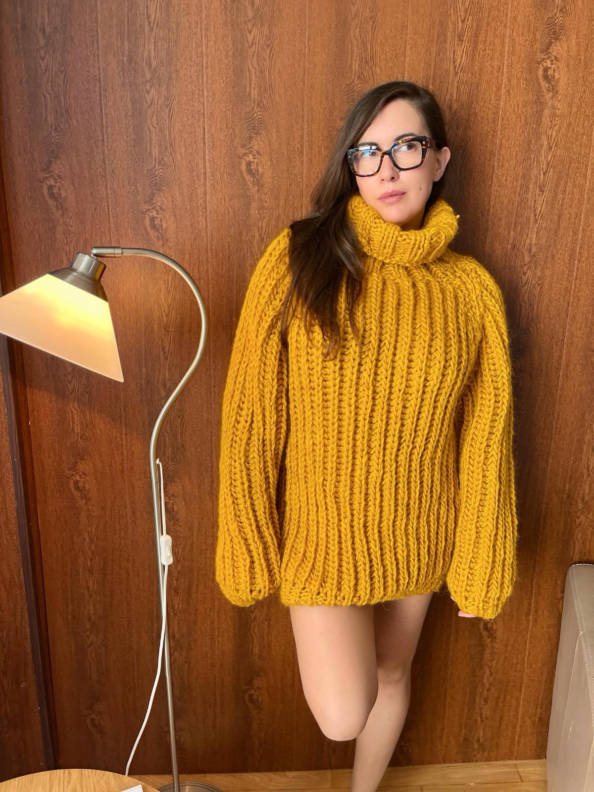 Woman wearing a thick mustard-yellow chunky knit turtleneck sweater against a wood wall.