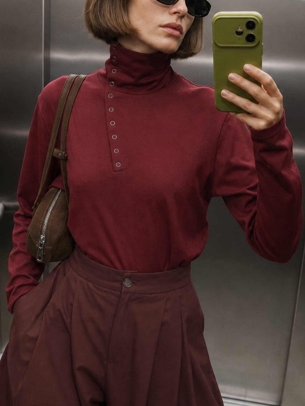 Woman in a maroon buttoned turtleneck top taking a mirror selfie in an elevator.
