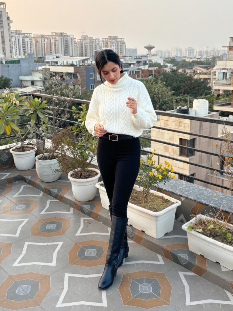 15 Long Boots Outfit Ideas for Cold Months Ahead 11 Woman in a white turtleneck sweater, black pants, and black knee-high boots standing on a rooftop.