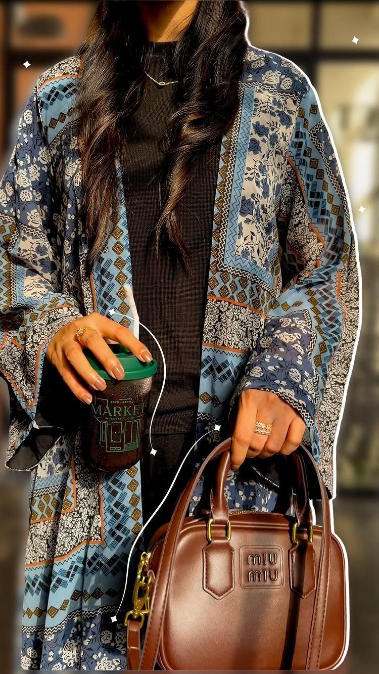 Person wearing a patterned blue bohemian long cardigan holding a coffee cup and leather handbag.