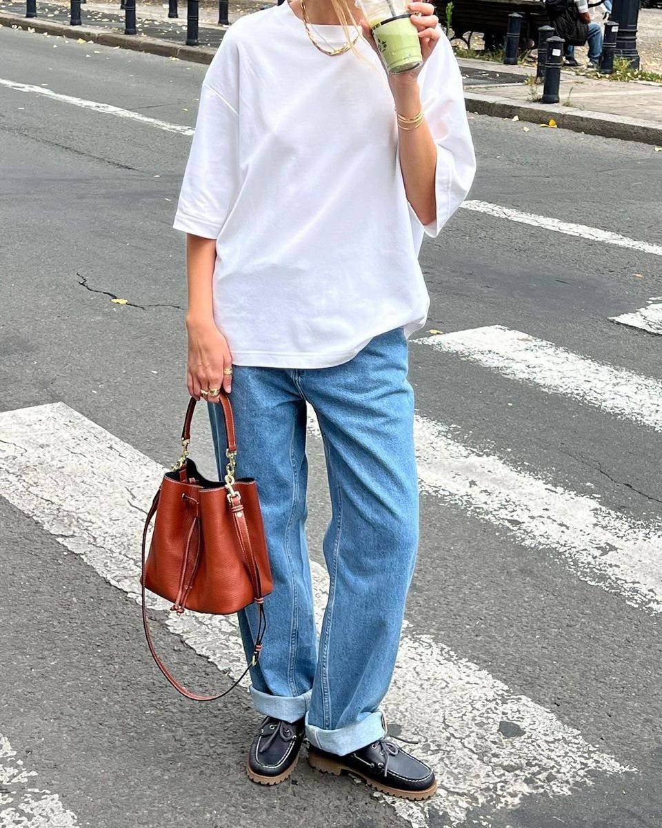 Person wearing an oversized white t-shirt, blue cuffed jeans, and black loafers while holding a brown handbag.