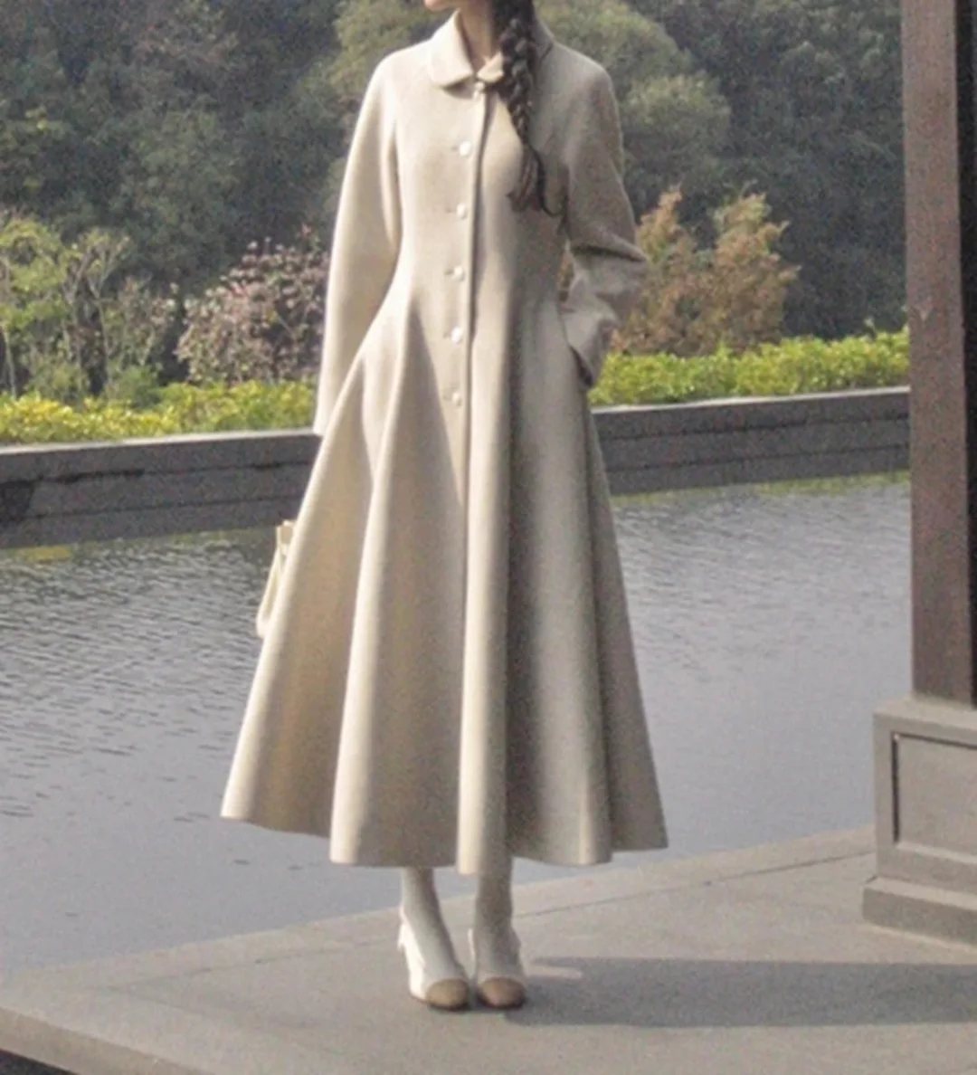 A woman wearing a long, cream-colored A-line dress coat standing near water.