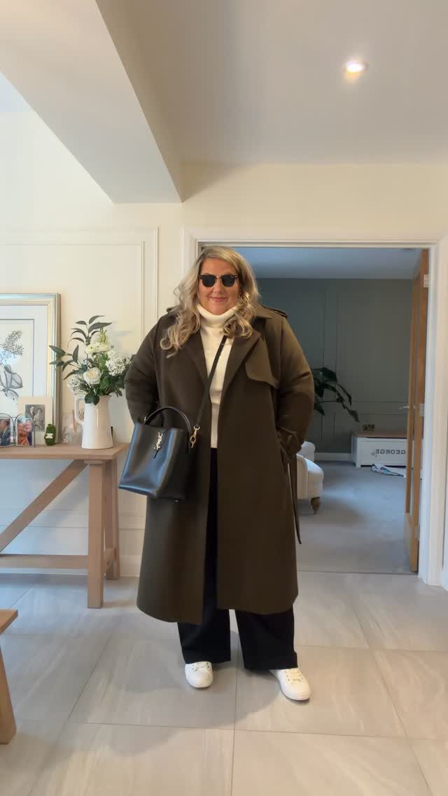 A blonde woman wearing a white turtleneck, olive green coat, and black wide-leg trousers standing indoors.